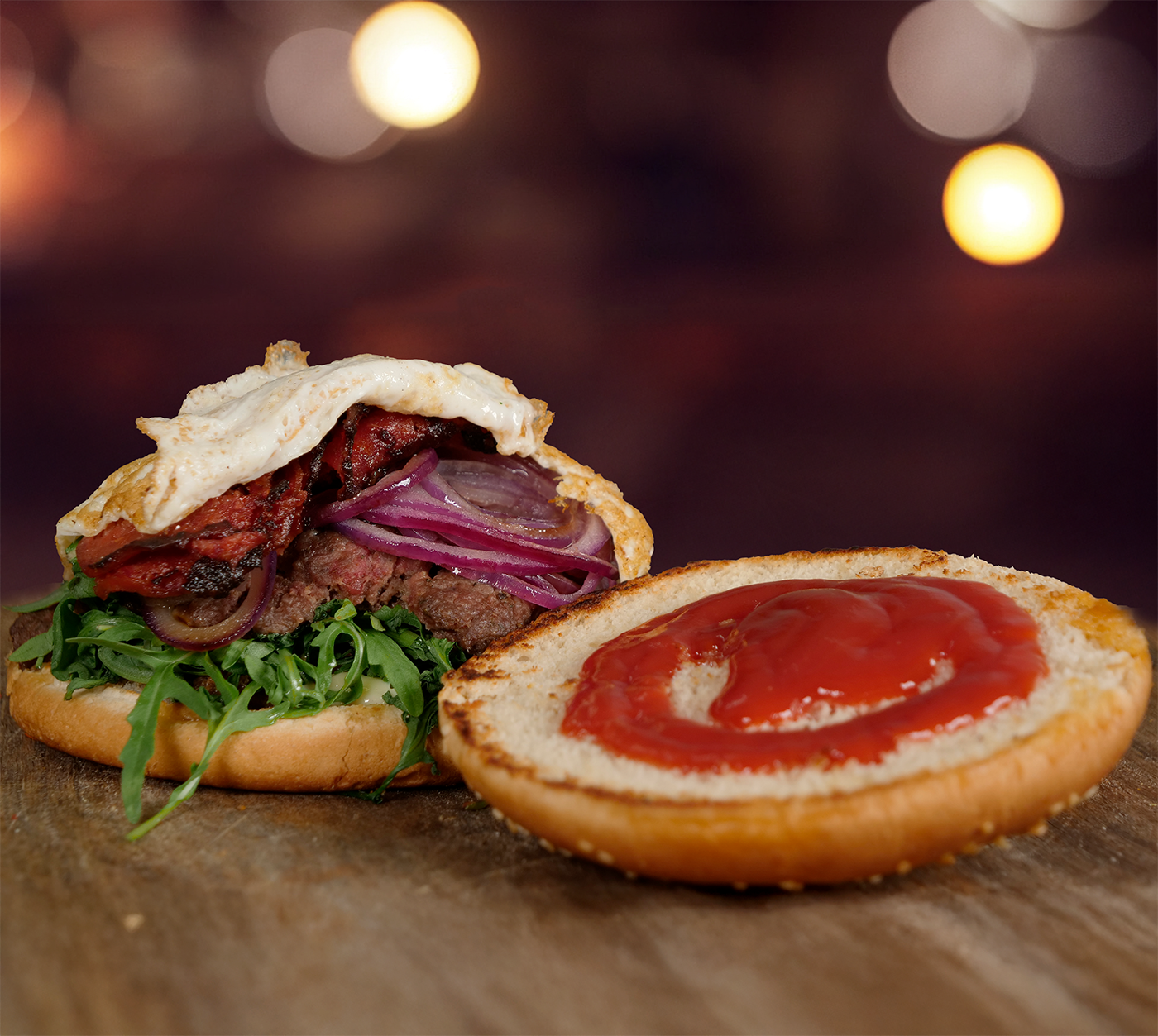 Seitliche Nahaufnahme vom Burger mit Ei, Rucola, roten Zwiebeln und Grillgemüse – saftig angerichtet auf einem rustikalen Holzbrett, Pommes im Hintergrund leicht verschwommen.