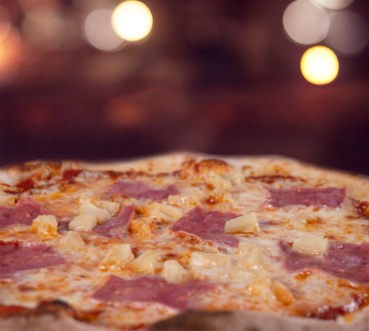 Pizza Hawaii seitlich fotografiert – geschmolzener Käse trifft auf fruchtige Ananasstücke und herzhaften Schinken, vor warmem, stimmungsvollem Hintergrund.