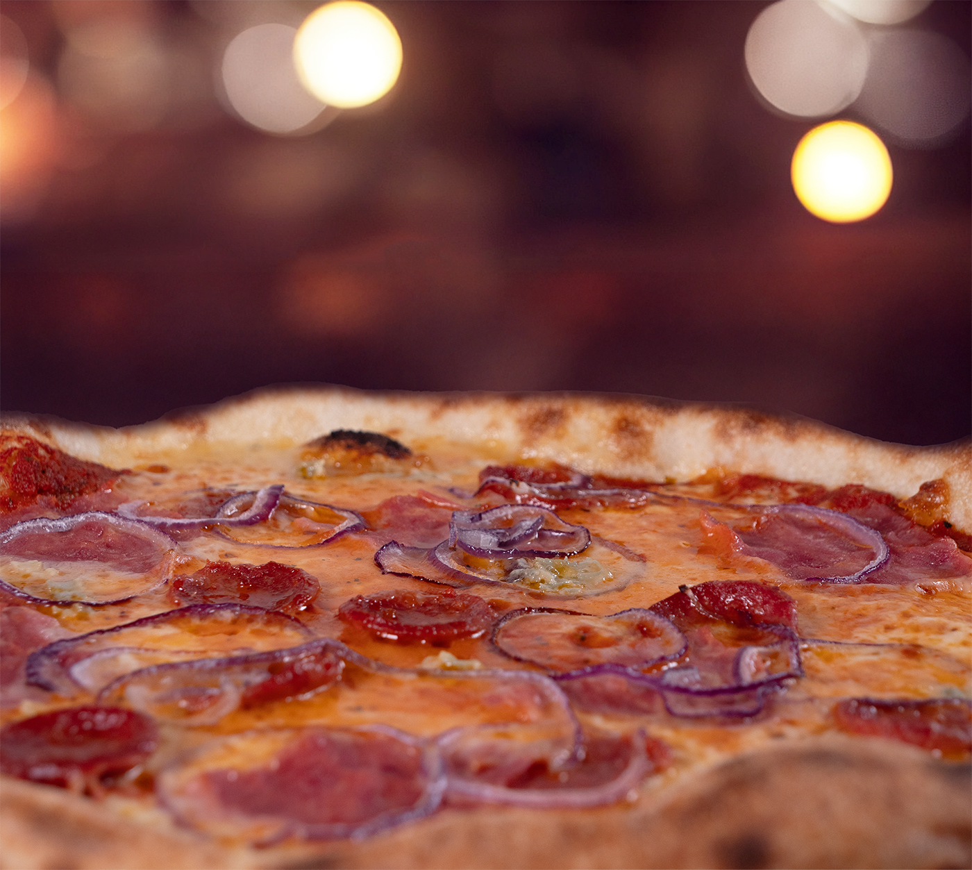 Pizza La Mana seitlich fotografiert – fluffiger Rand, cremiger Käse, herzhafte Salami und Schinken mit Zwiebeln, stimmungsvolles Licht im Hintergrund.