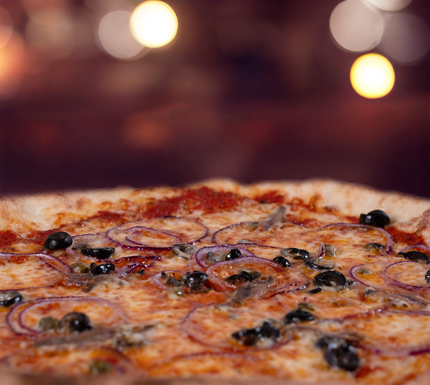 Pizza Siciliana seitlich fotografiert – luftiger Rand, intensiver Belag mit Oliven, Sardellen, Zwiebeln und Kapern, warmes Licht im Hintergrund.