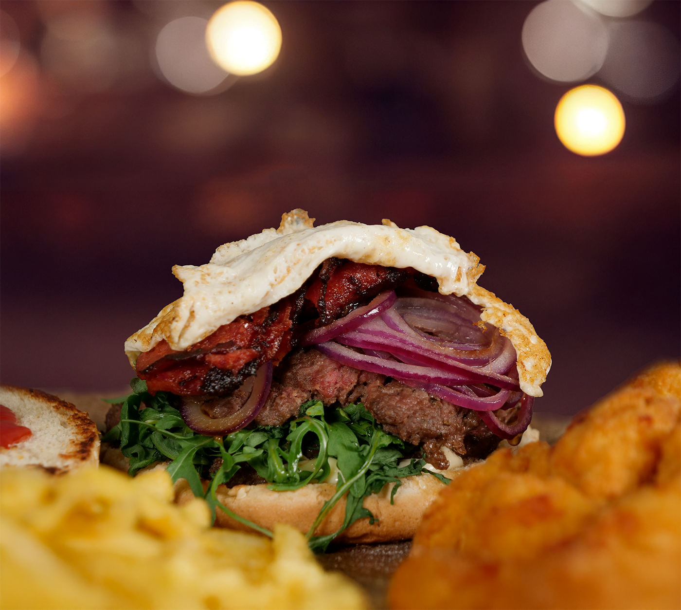 Saftiger Burger mit gebratenem Ei, roten Zwiebeln und Rucola – perfekt geschichtet, daneben Pommes und Nuggets im Anschnitt sichtbar.