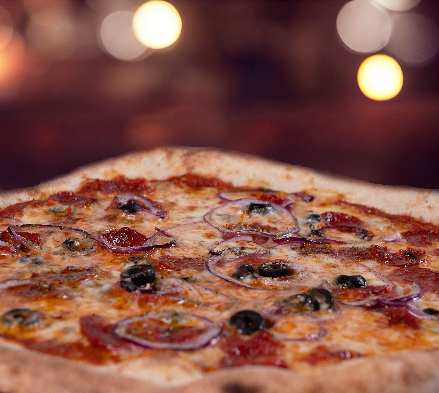 Pizza Calabrese seitlich fotografiert – knuspriger Rand, scharfe Salami, Oliven und Zwiebeln auf goldbraunem Käse, weiches Licht im Hintergrund.