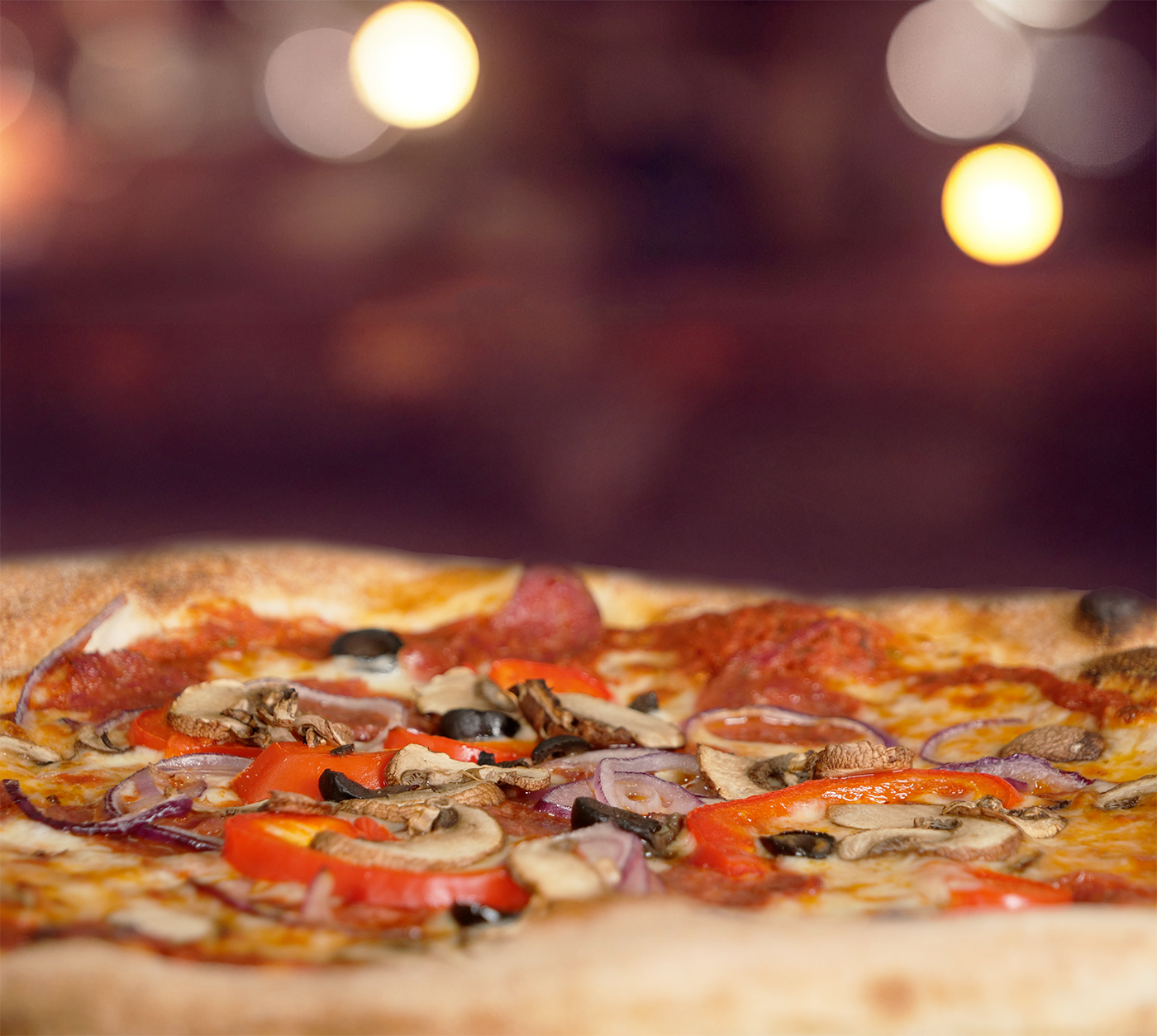 Pizza Capricciosa seitlich fotografiert – geschmolzener Käse trifft auf herzhafte Zutaten, stimmungsvoll beleuchtet mit warmem Bokeh-Effekt im Hintergrund.