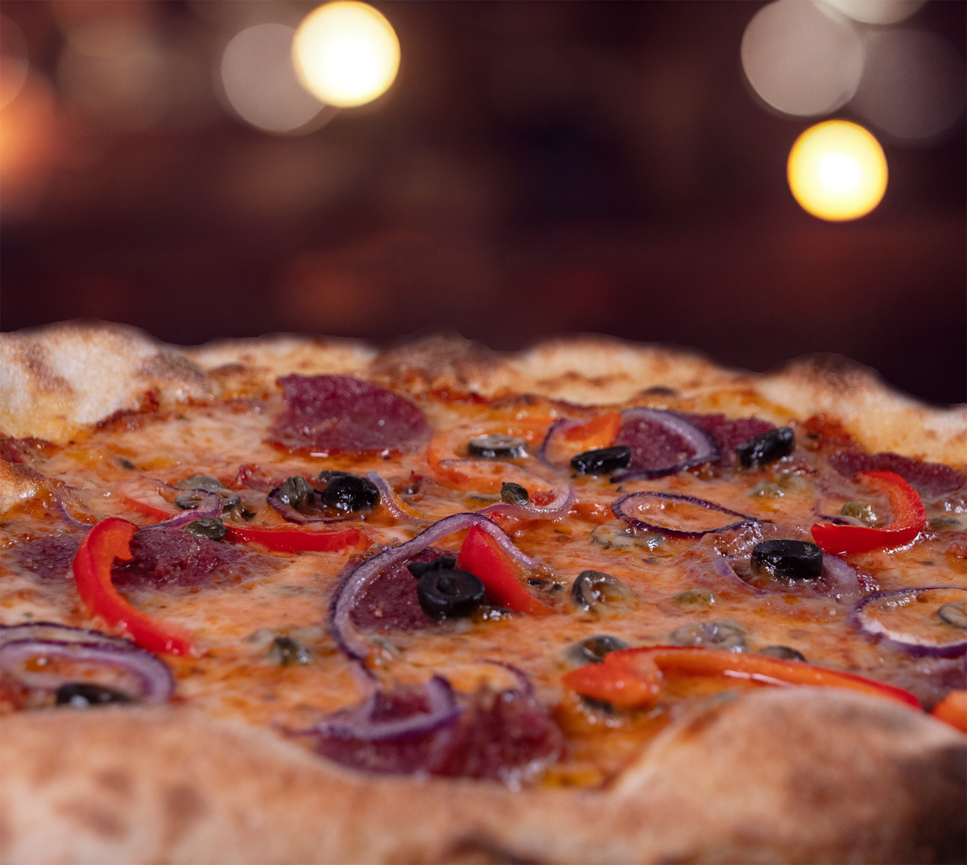 Pizza Diavolo seitlich fotografiert – fluffiger Rand, würziger Belag mit Salami, Peperoni, Oliven und Zwiebeln, leicht geschmolzenem Käse, stimmungsvolle Hintergrundbeleuchtung.