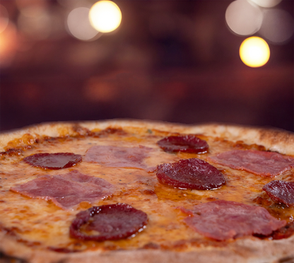 Pizza Fantasia seitlich fotografiert – luftiger Rand, zart geschmolzener Käse, herzhafte Salamischeiben und Schinken, stimmungsvolle Beleuchtung im Hintergrund.