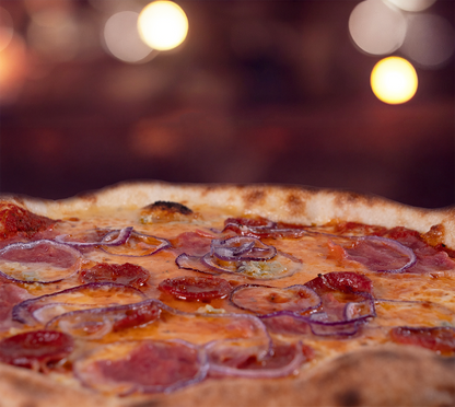 Pizza La Mana seitlich fotografiert – fluffiger Rand, cremiger Käse, herzhafte Salami und Schinken mit Zwiebeln, stimmungsvolles Licht im Hintergrund.