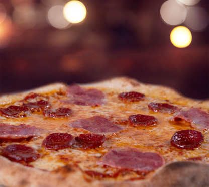 Pizza Lina seitlich fotografiert – knuspriger Rand, kräftig belegte Oberfläche mit Salami, Schinken und Gorgonzola, warm ausgeleuchtet im Hintergrund.