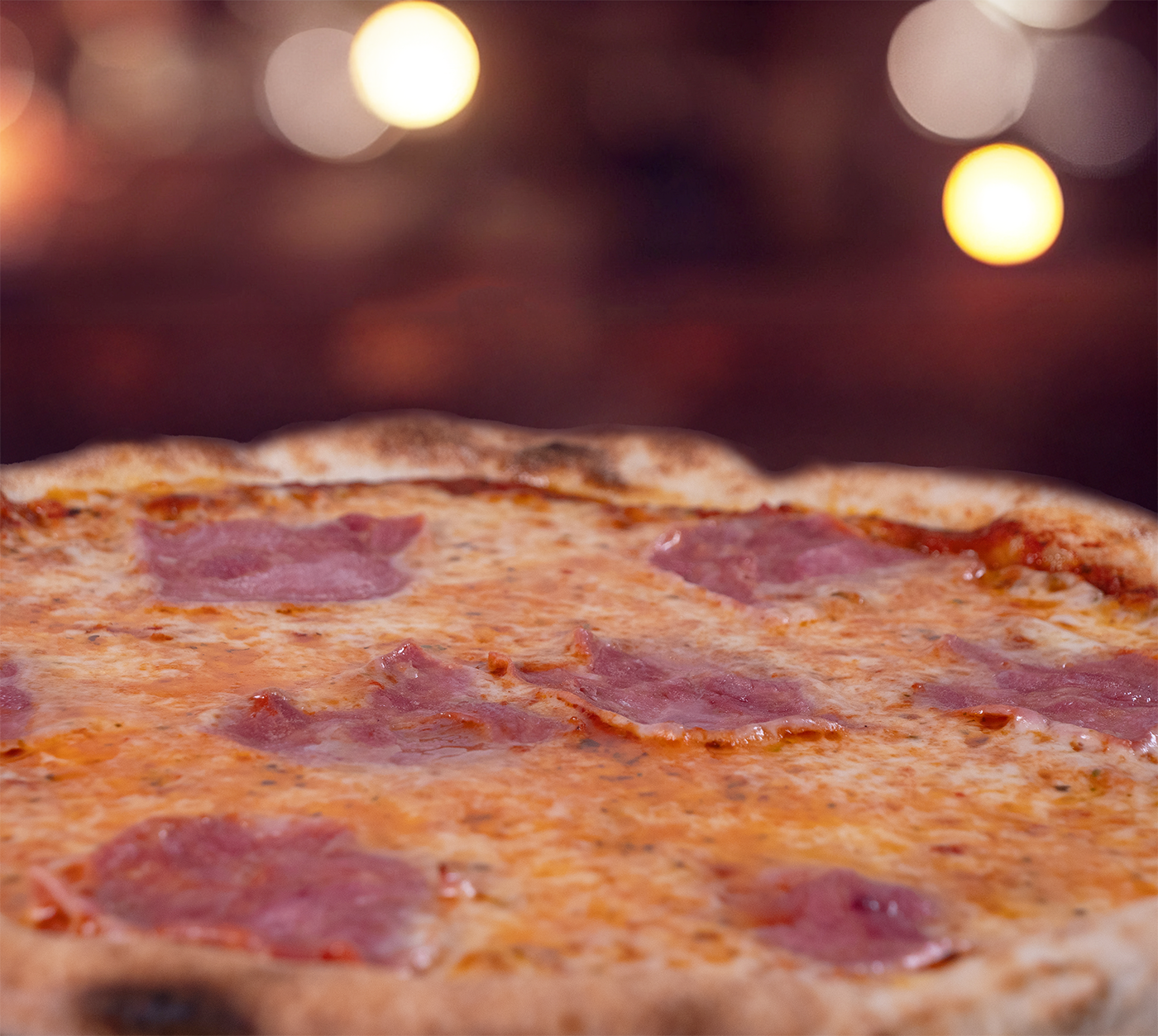 Pizza Prosciutto seitlich fotografiert – fluffiger, goldbrauner Rand, saftiger Schinken auf geschmolzenem Käse, warm ausgeleuchtet für echten Genussmoment.