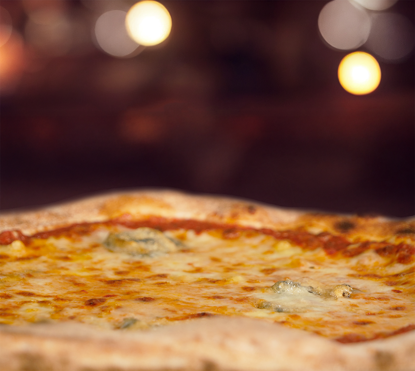 Pizza Quattro Formaggi seitlich fotografiert – geschmolzener Käsemix auf fluffigem Rand, serviert im warmen Licht mit Bokeh im Hintergrund.