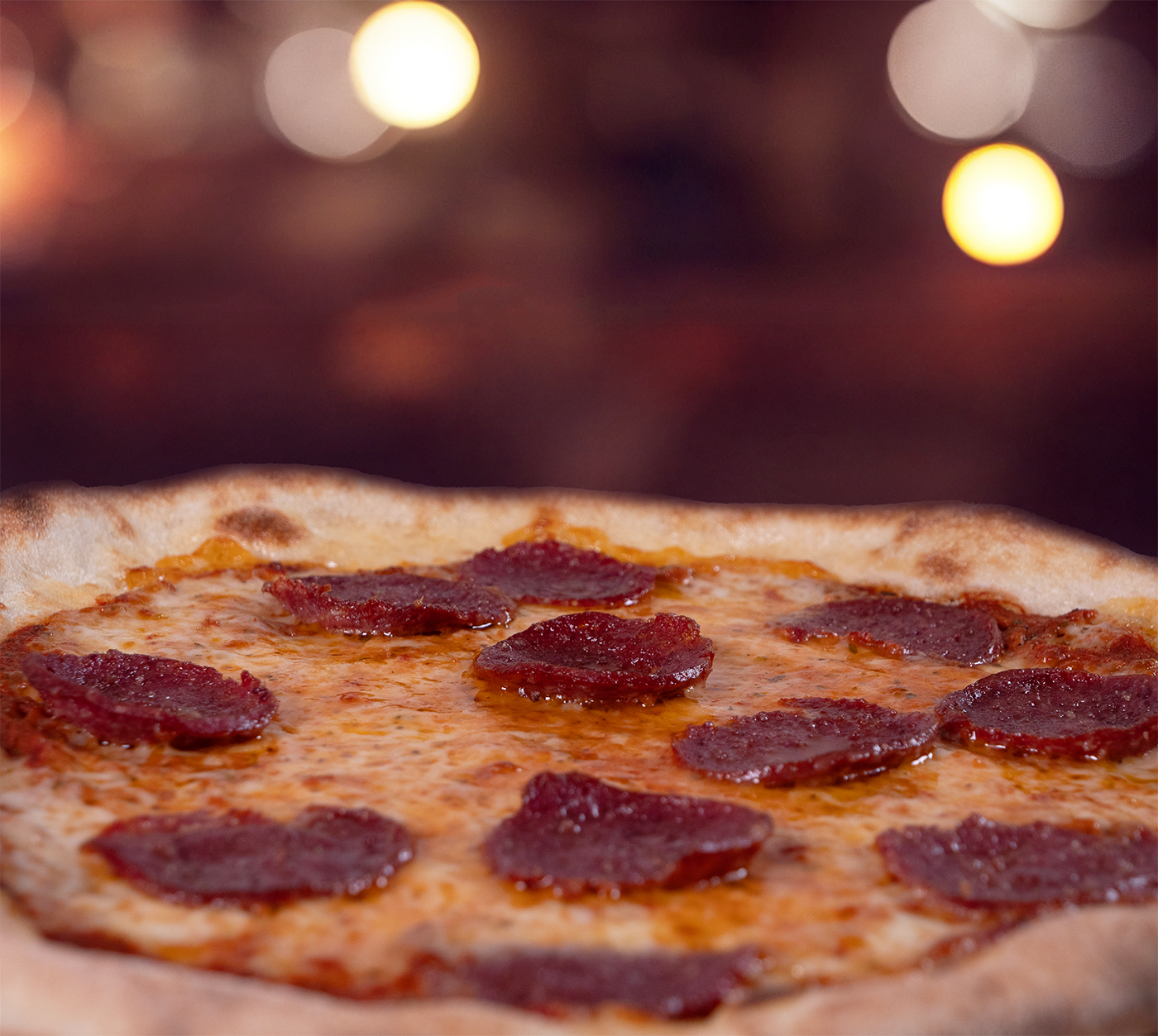 Pizza Salami seitlich fotografiert – luftiger Rand, geschmolzener Käse und Salamischeiben im Fokus, serviert auf Holzbrett, warme Food-Szene.