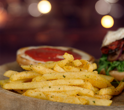 Nahaufnahme von gewürzten Pommes mit feinem Kräutermantel – im Hintergrund leicht verschwommen der Burger mit Ketchup und Rucola.